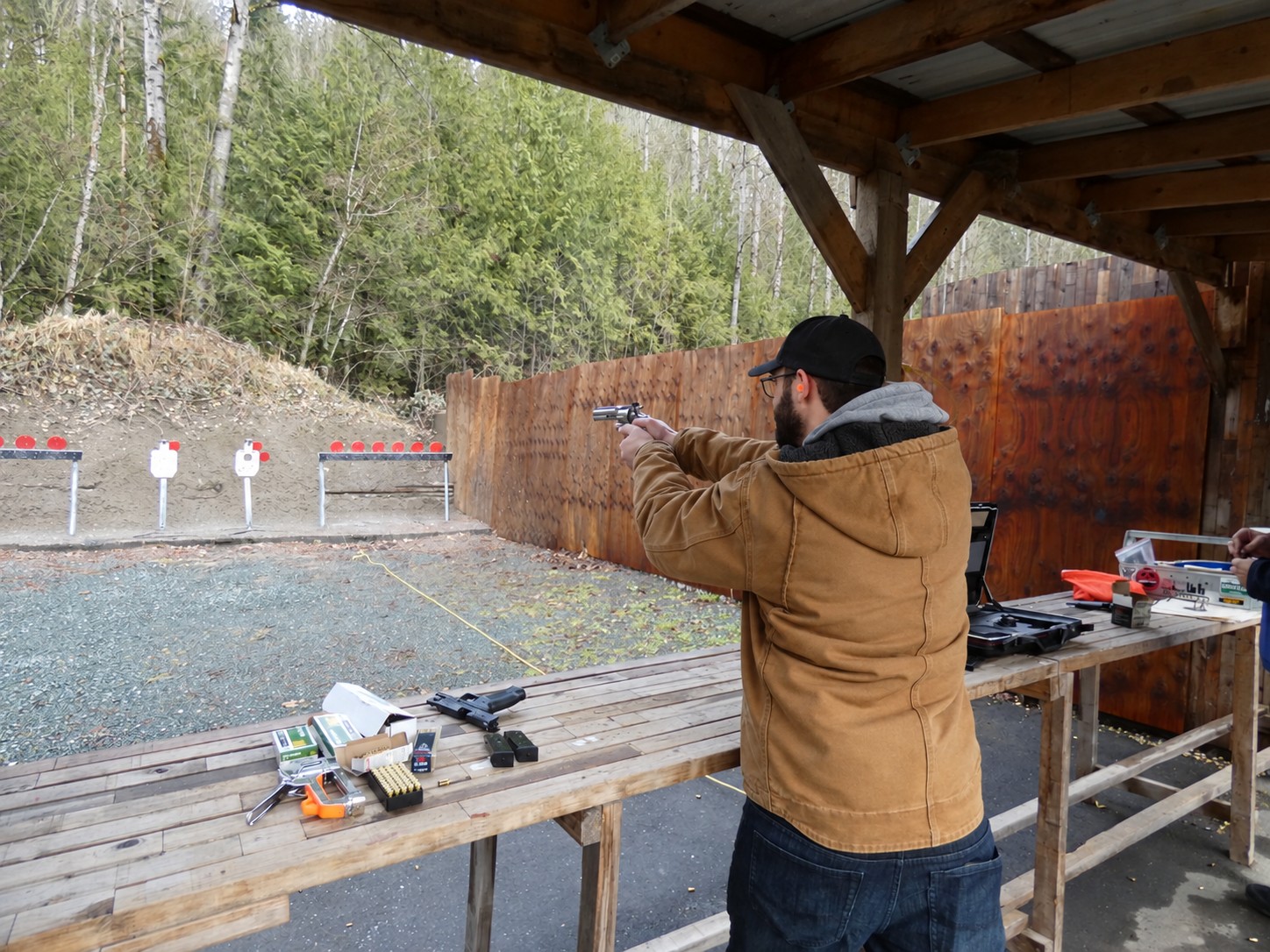 A man wearing eye and hearing protection aiming a handgun toward targets at a wooded outdoor range.